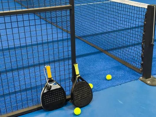 Dos paletas negras apoyadas en la red de una cancha azul, con pelotas amarillas en el piso